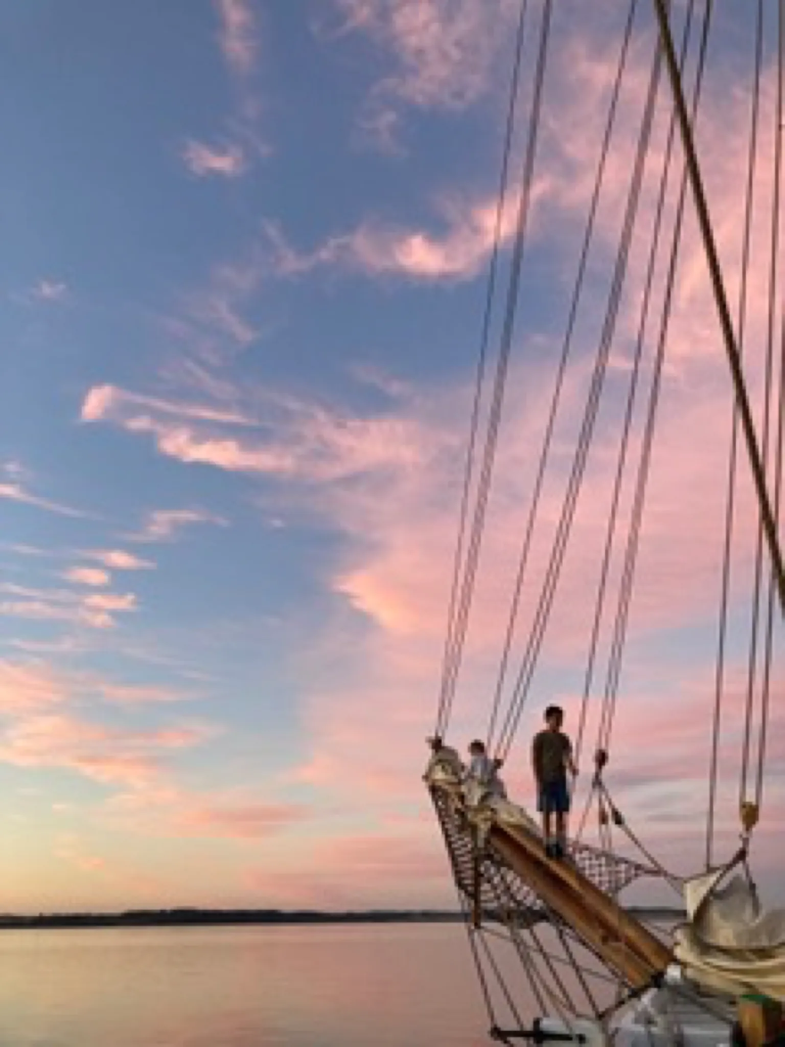 Two children stand on the bowsprit under a pink evening sky
